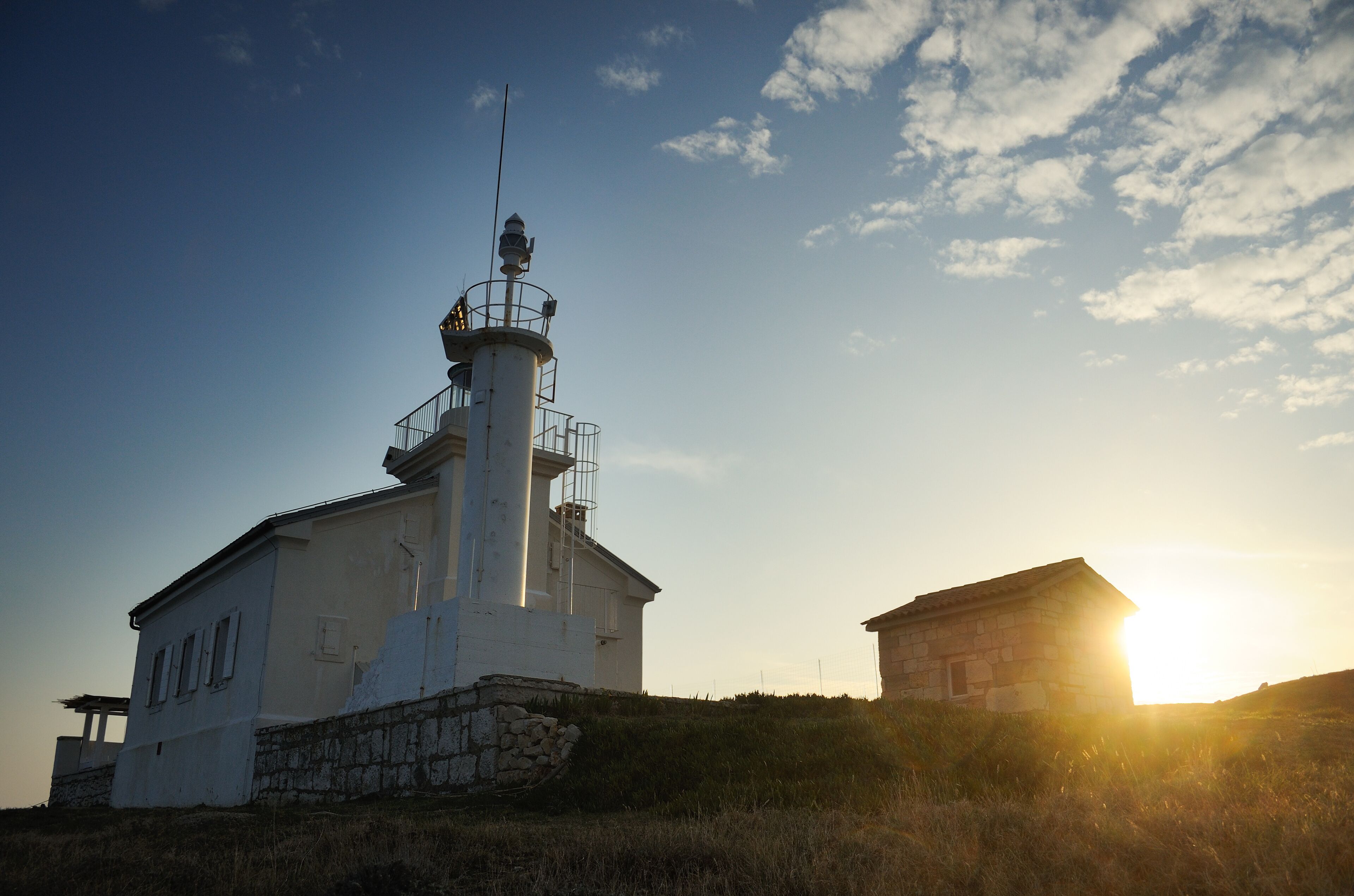 A lighthouse with a stone base and a white upper structure, set against a clear sky with the sun low on the horizon.