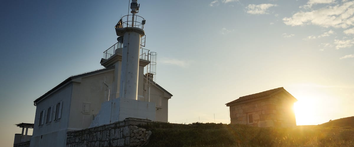 A lighthouse with a stone base and a white upper structure, set against a clear sky with the sun low on the horizon.