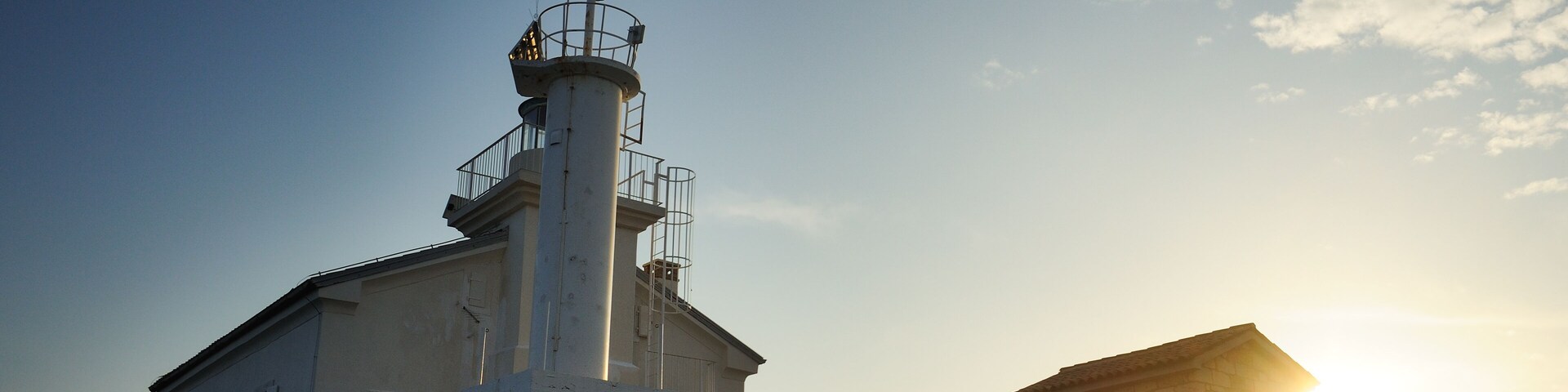 A lighthouse with a stone base and a white upper structure, set against a clear sky with the sun low on the horizon.