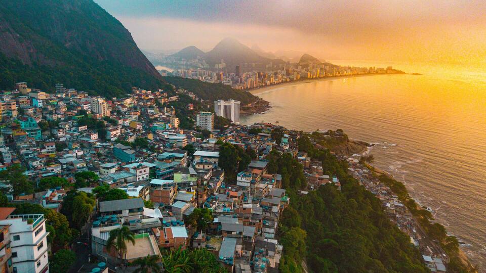 Paisagem costeira urbana com edifícios densos, uma montanha imponente e um pôr do sol sobre o oceano.