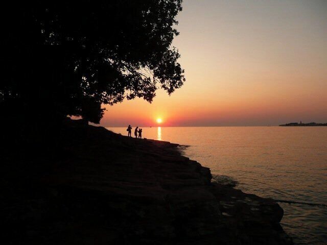 A sunset over a calm sea with silhouettes of people on a rocky shore.