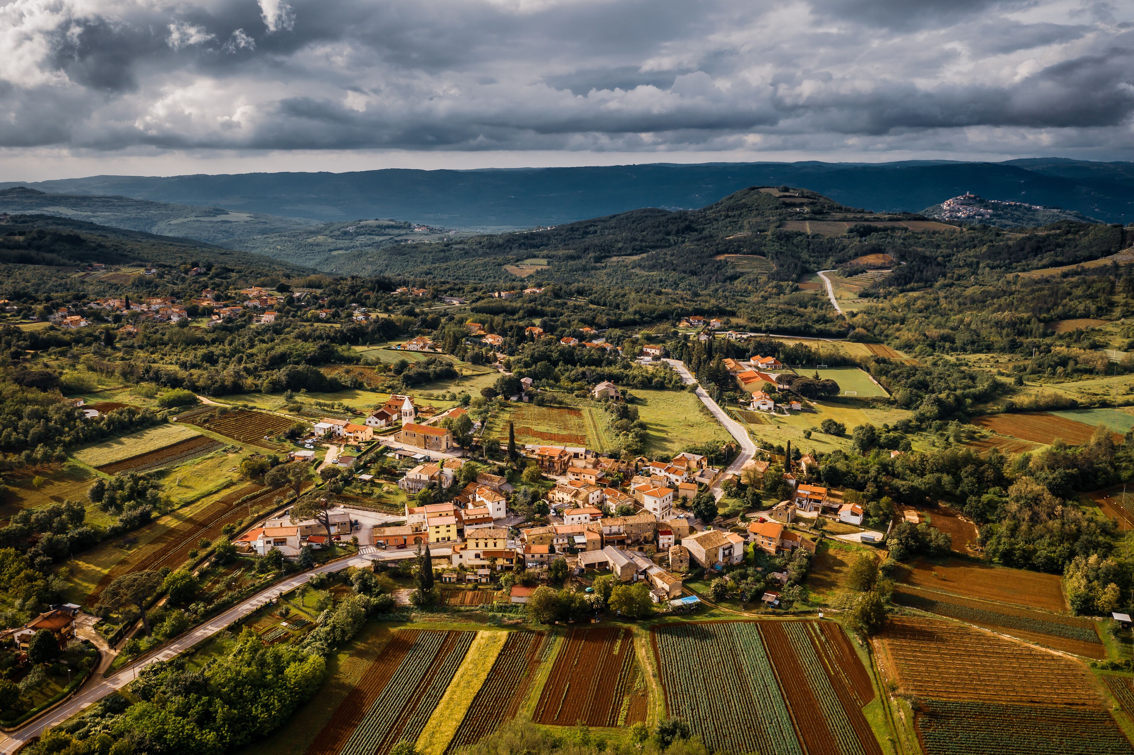 An aerial view of a village with terraced fields, a winding road, and rolling hills in the background.