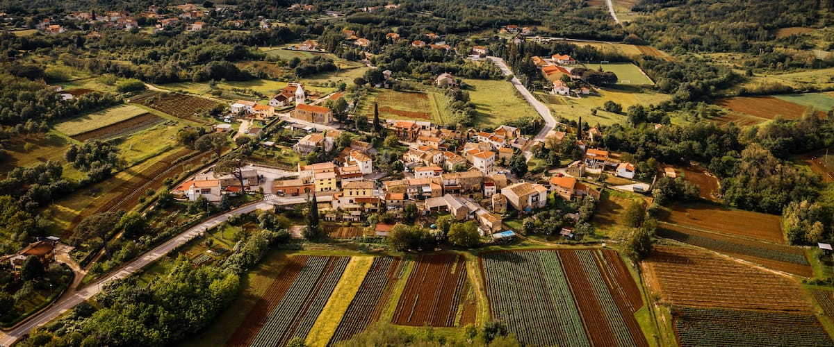 An aerial view of a village with terraced fields, a winding road, and rolling hills in the background.