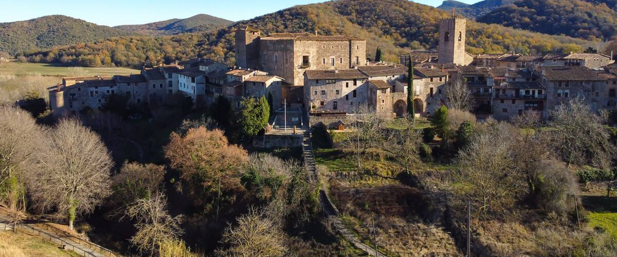 A medieval village with stone buildings and a prominent tower, surrounded by hills and trees.