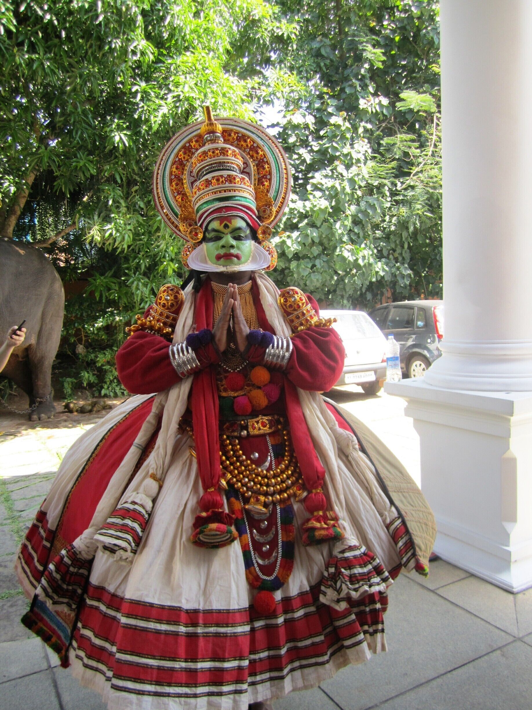 A person in traditional attire with elaborate headgear, standing outdoors with trees and a statue in the background.