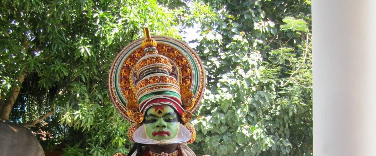 A person in traditional attire with elaborate headgear, standing outdoors with trees and a statue in the background.