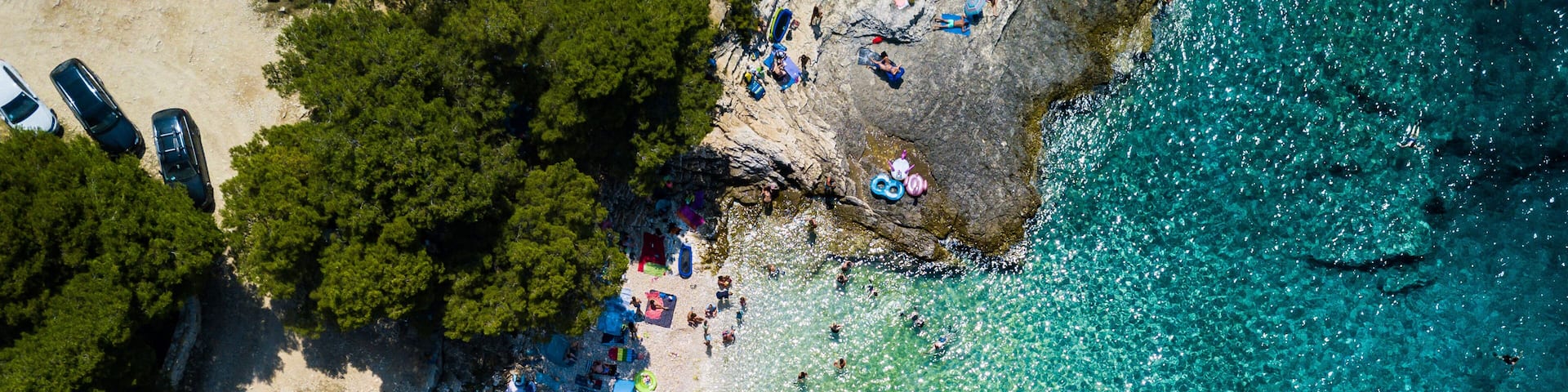 An aerial view of a beach with clear turquoise water, a sandy shore, and several parked cars.
