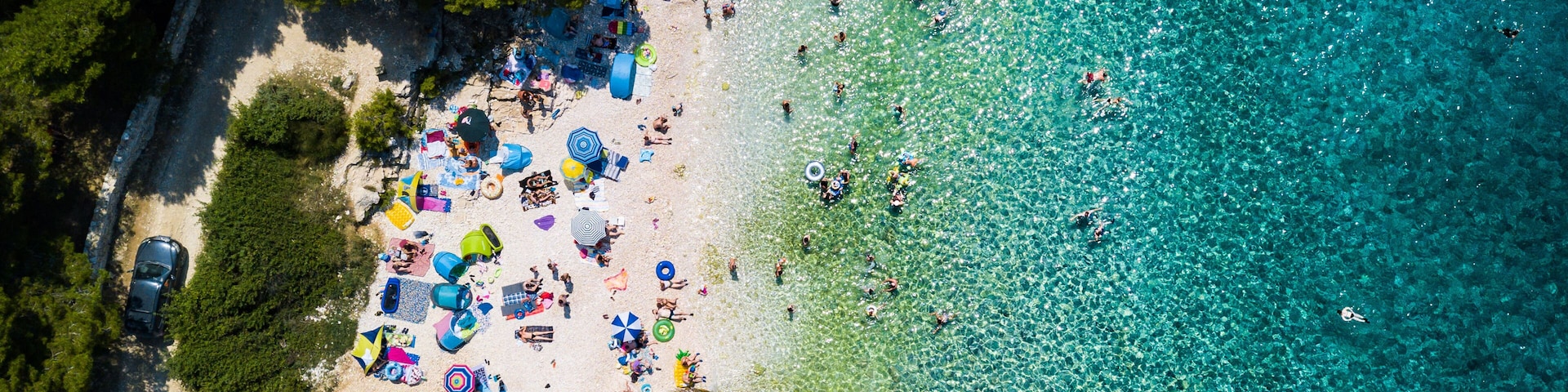 Ein Luftbild einer Strandbucht mit klarem tĂŒrkisblauem Wasser und einem Sandstrand, darunter einige parkende Autos.
