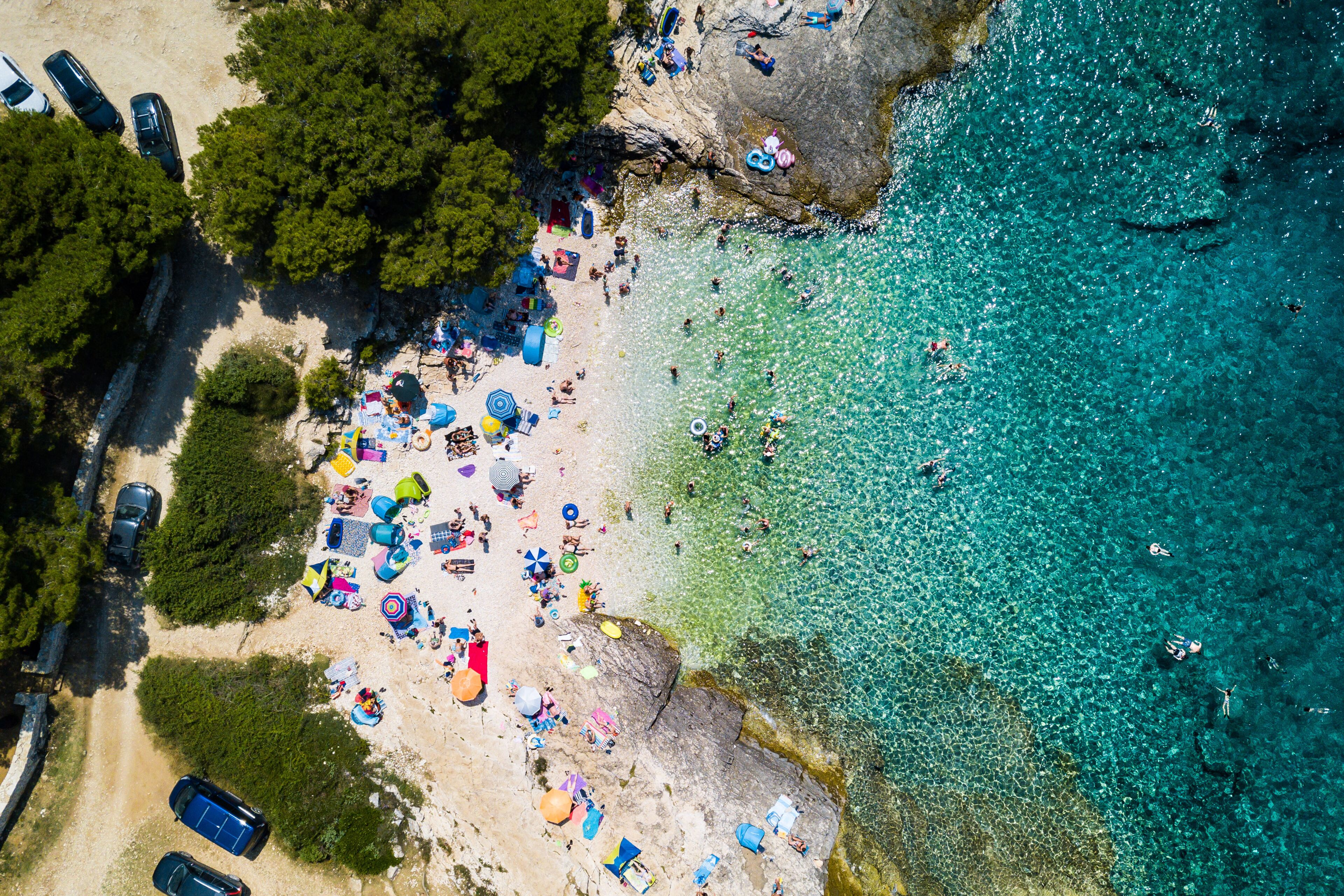 An aerial view of a beach with clear turquoise water, a sandy shore, and several parked cars.