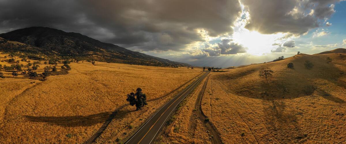 A railway track through a dry, hilly landscape with a mountain in the background and a partly cloudy sky.
