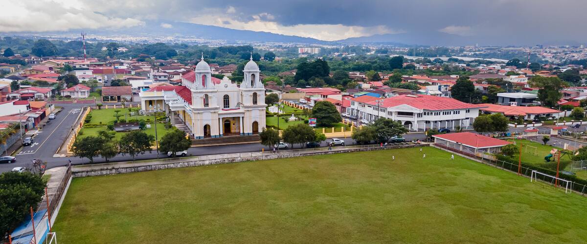 A large white church with a bell tower, surrounded by a green field and residential buildings.