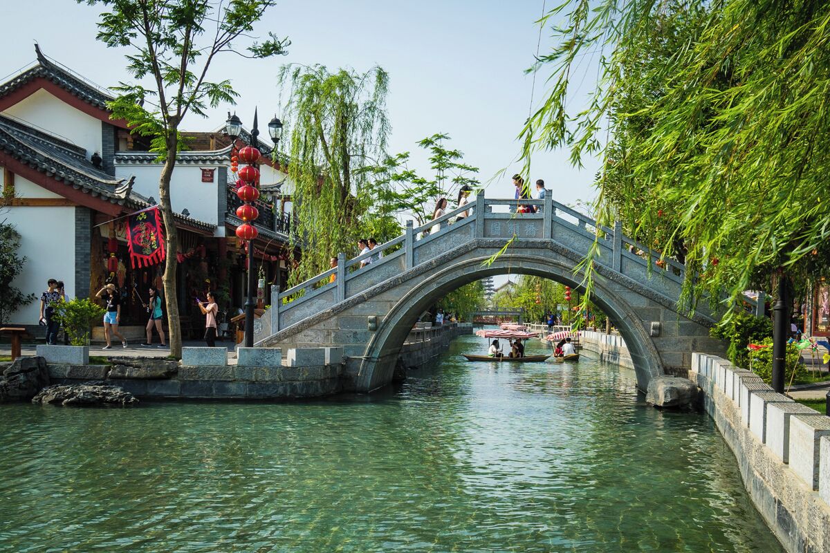 A stone bridge over a river with traditional buildings and red lanterns on the left.