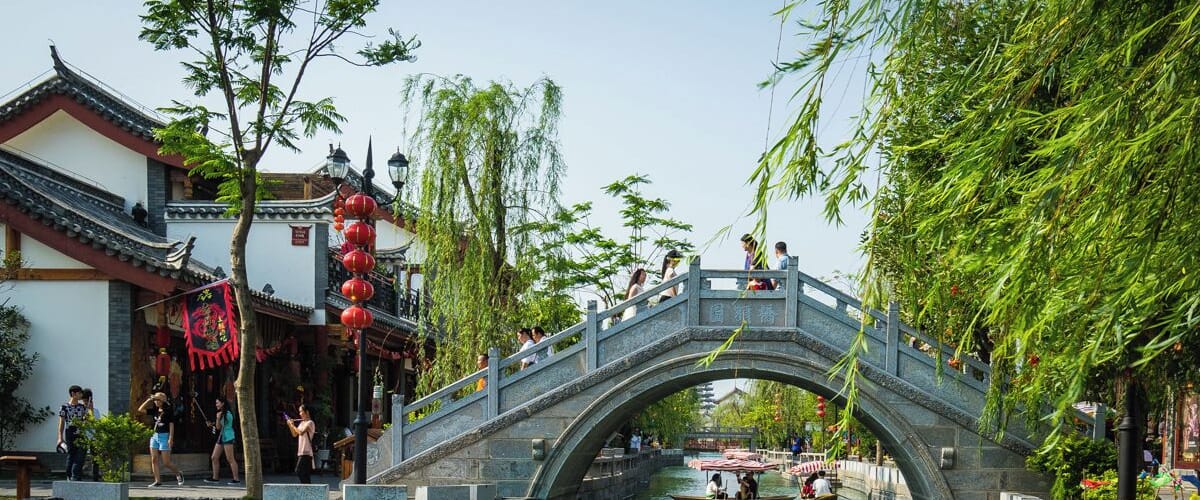 A stone bridge over a river with traditional buildings and red lanterns on the left.
