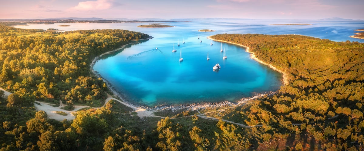 A coastal landscape with a turquoise bay, surrounded by dense forests, and several sailboats anchored in the water.