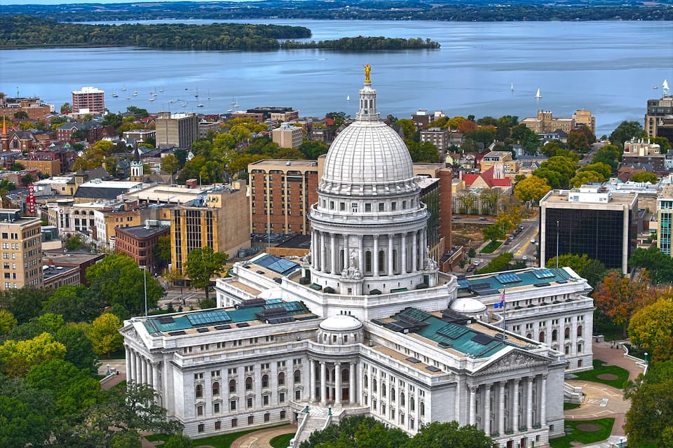A large domed building with a statue on top, surrounded by trees and buildings, with a body of water in the background.