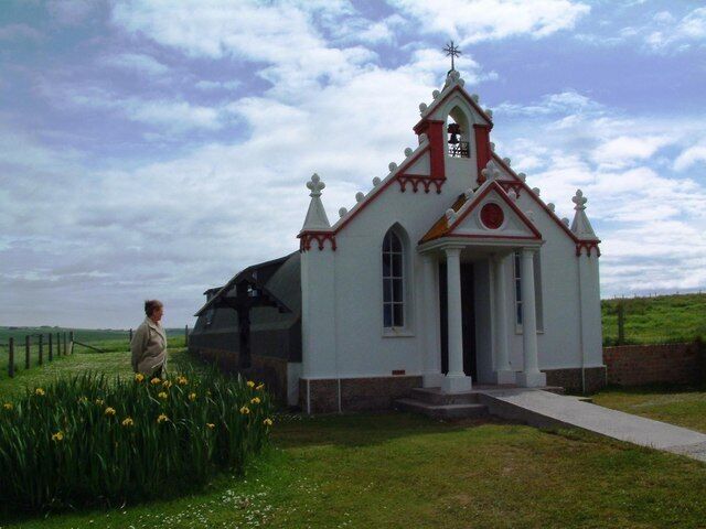 A small white church with red trim and a steeple, surrounded by green grass and yellow flowers.