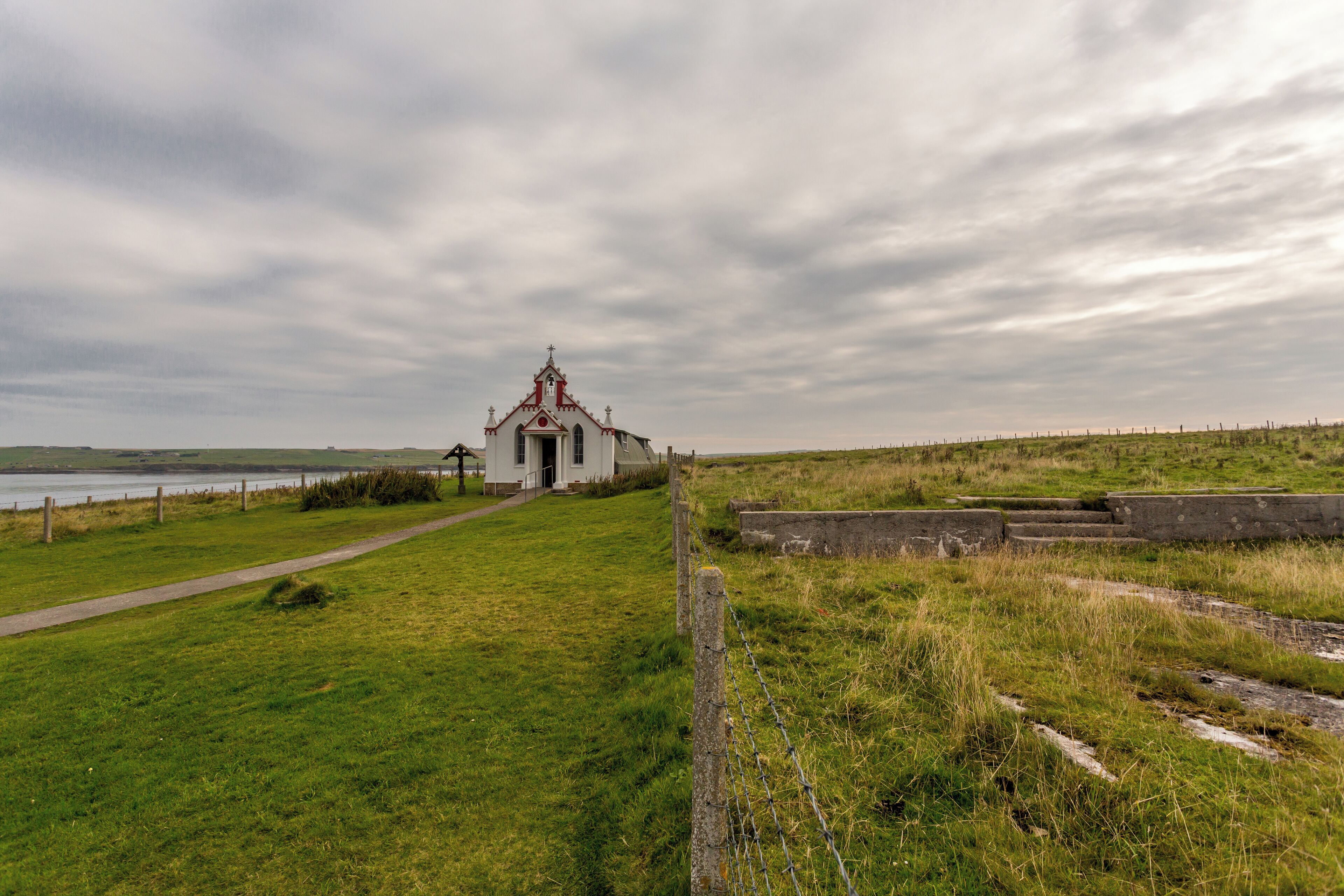 A small white church with a red roof and a cross on top, surrounded by grassy fields and a body of water in the background.
