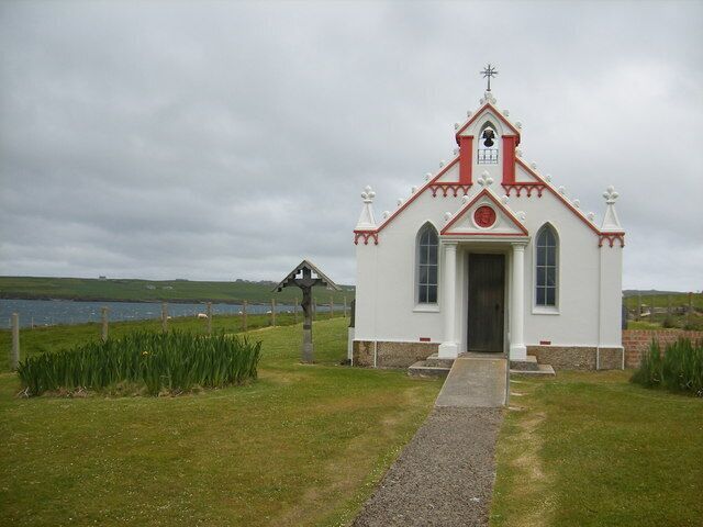A small white church with a red roof and a weather vane, situated by a body of water.