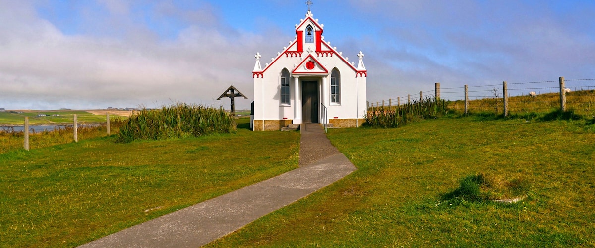 A small white church with a red roof and a cross on top, surrounded by a grassy field and a fence.