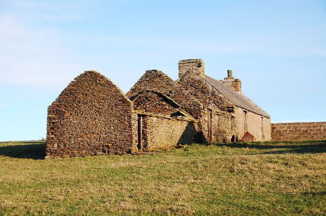 A stone structure with multiple roofs and a brick wall in a grassy field.
