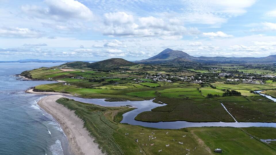 A coastal landscape with a beach, green fields, a river, and a mountain in the background.