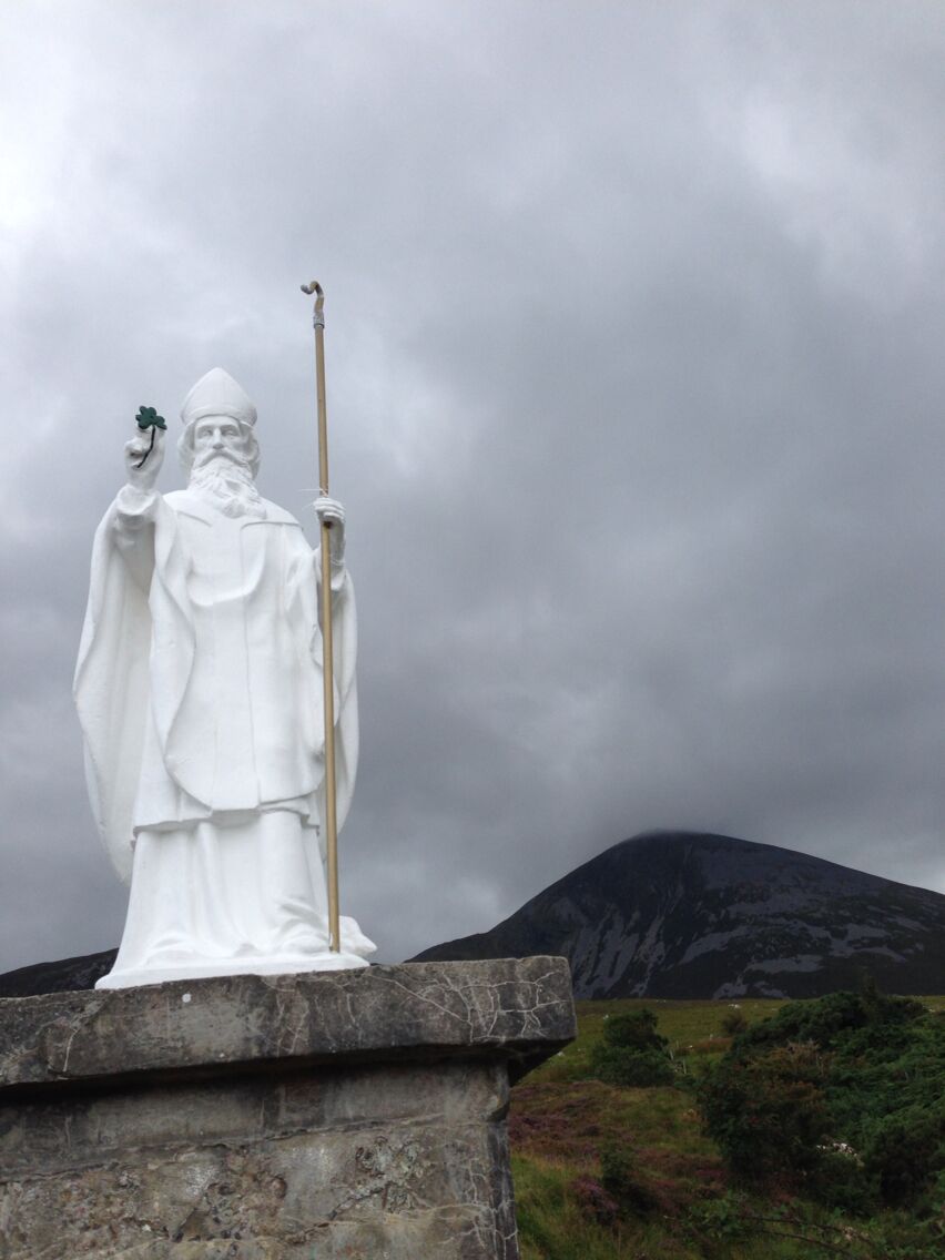 A white statue of a figure holding a staff and a shamrock, set against a cloudy sky with a mountain in the background.