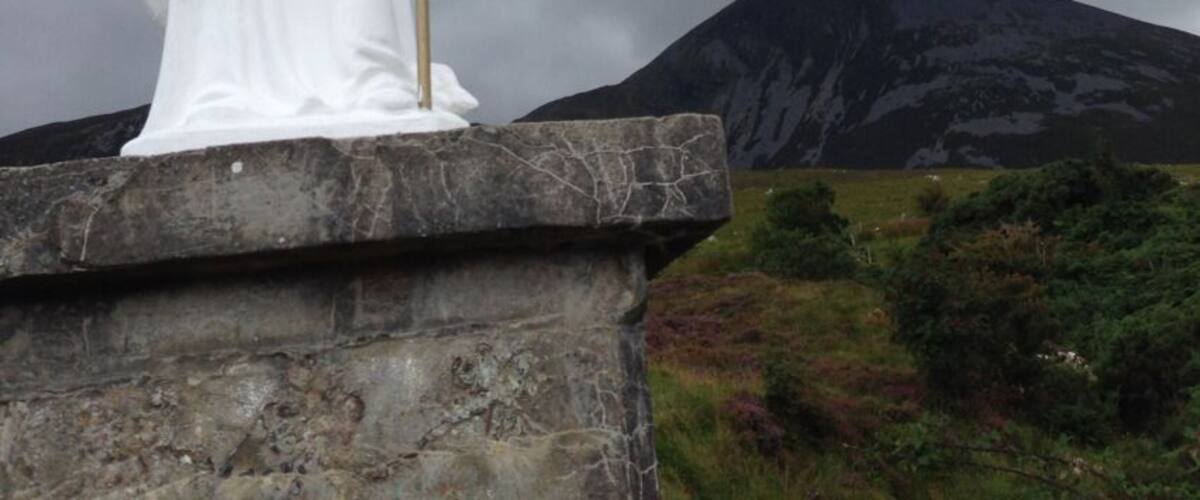 A white statue of a figure holding a staff and a shamrock, set against a cloudy sky with a mountain in the background.