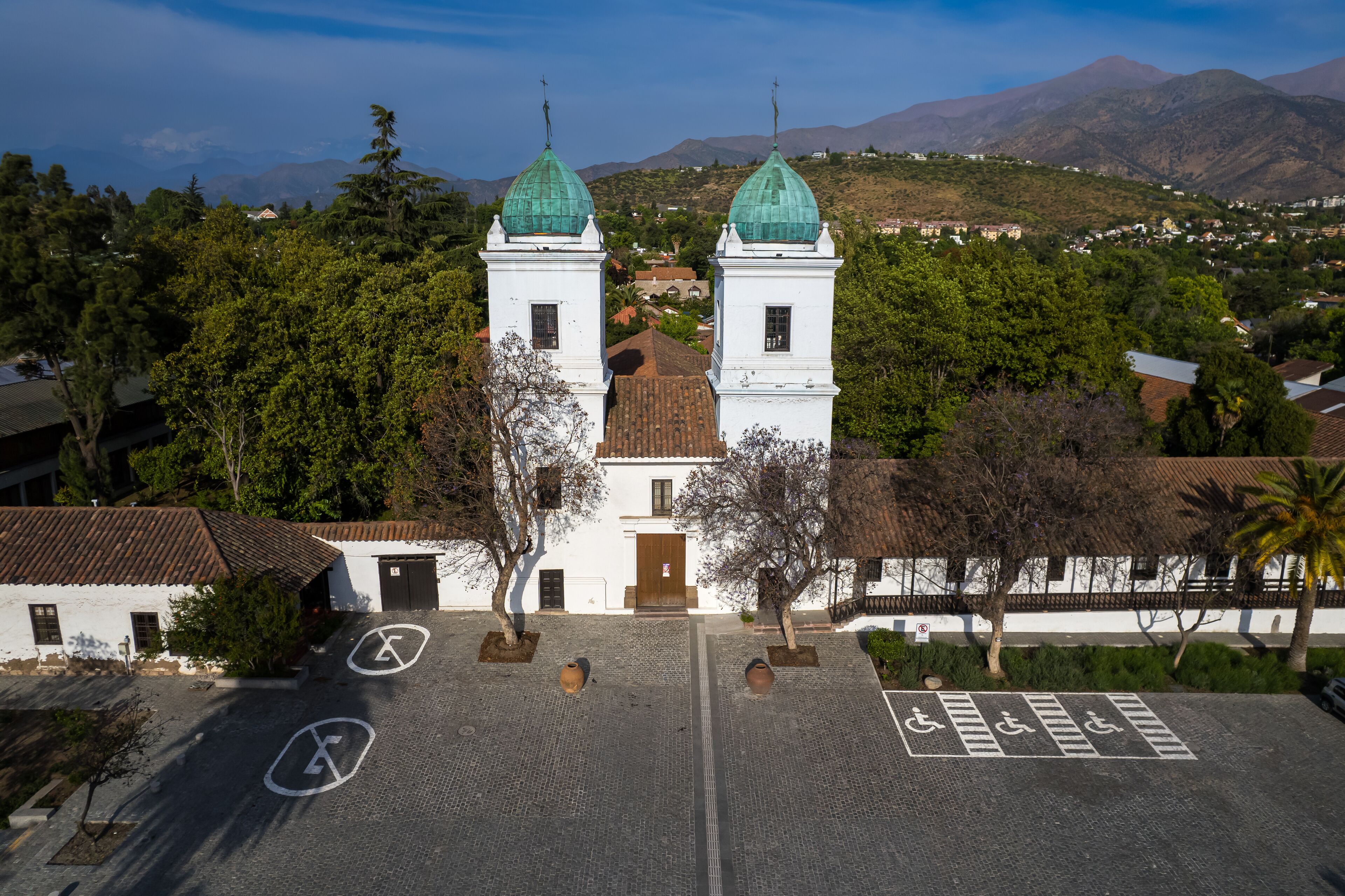 A white church with green domes, a paved courtyard, and a mountainous landscape in the background.