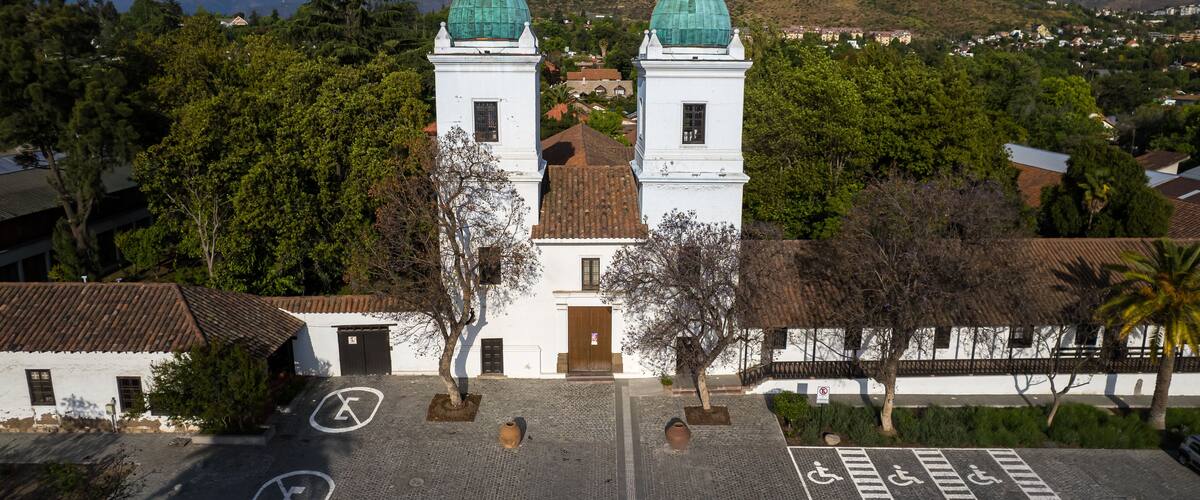 A white church with green domes, a paved courtyard, and a mountainous landscape in the background.