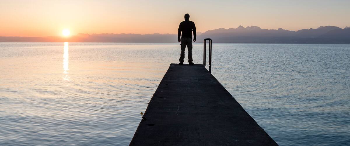 A person standing on a pier at sunset with mountains in the background.
