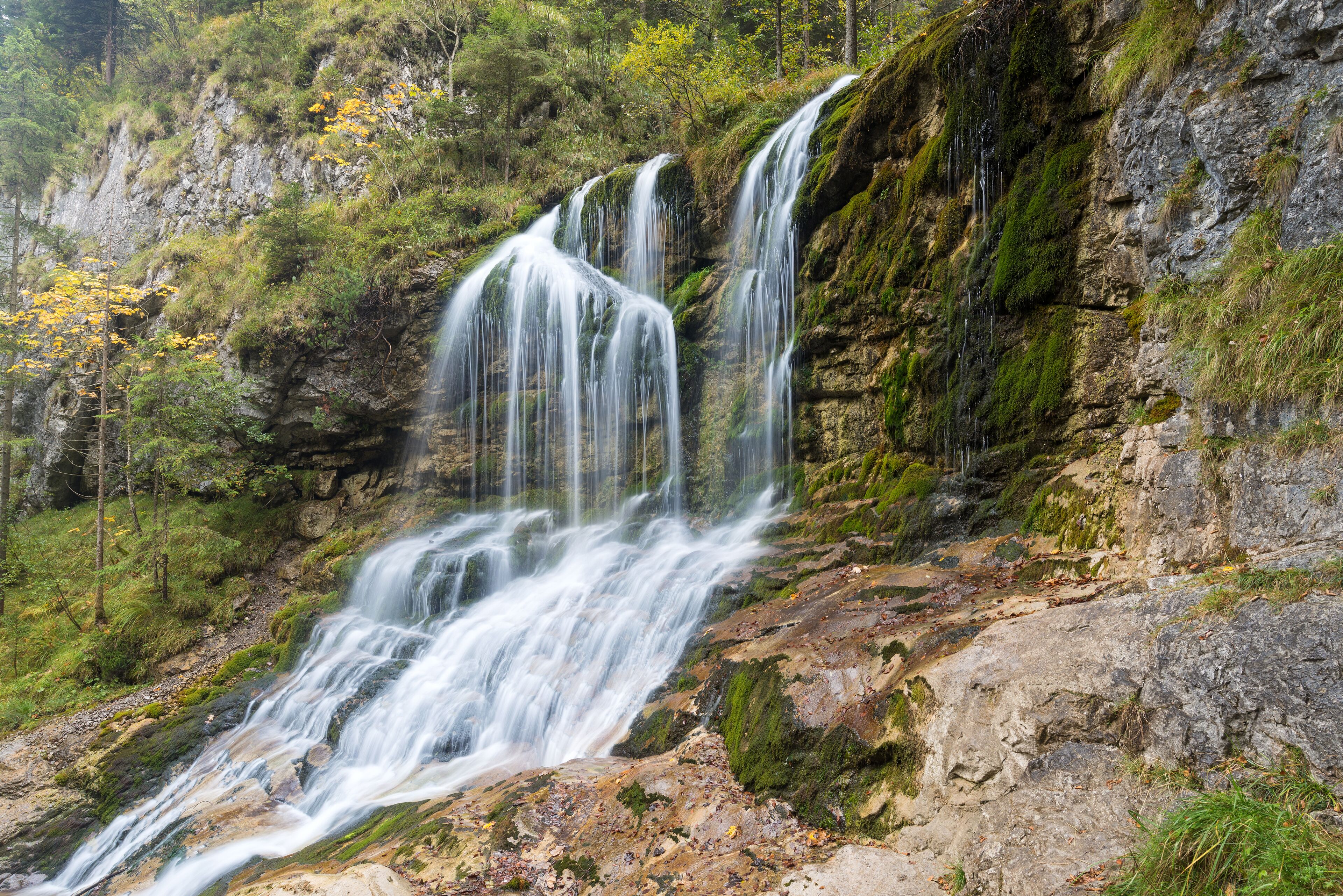 A multi-tiered waterfall cascading down a rocky cliff surrounded by lush greenery.