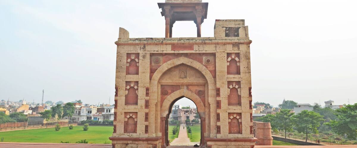 An ancient archway with intricate carvings and a dome on top, leading into a cityscape.