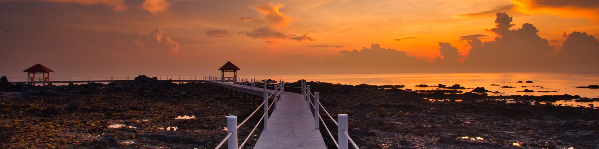 Un sendero de concreto que lleva a un pabellón sobre una costa rocosa al atardecer.
