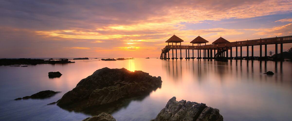A sunset over a calm sea with a pier and thatched-roof structures.