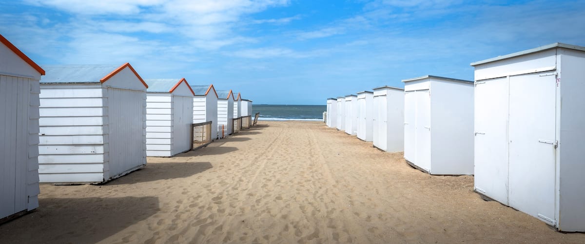 A row of white beach huts with orange roofs on a sandy beach, leading to the ocean.
