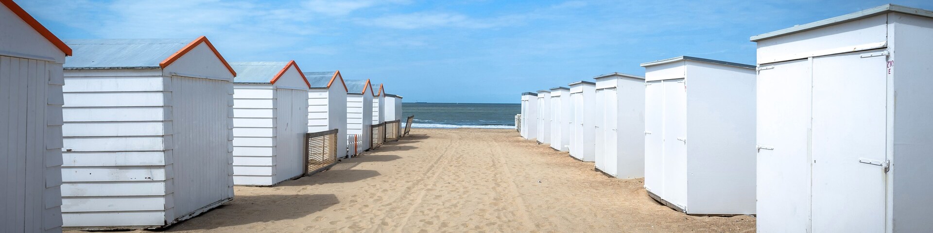A row of white beach huts with orange roofs on a sandy beach, leading to the ocean.