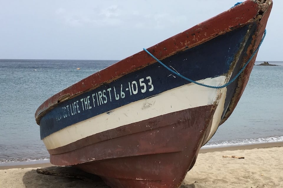 A weathered boat with a blue and white stripe and the text "LIFE THE FIRST L-1053" on its side, resting on a sandy beach with the ocean in the background.