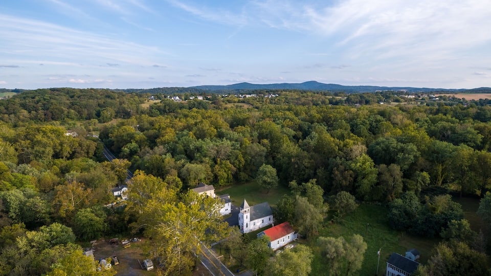 A lush green landscape with a white church surrounded by dense trees and rolling hills in the background.