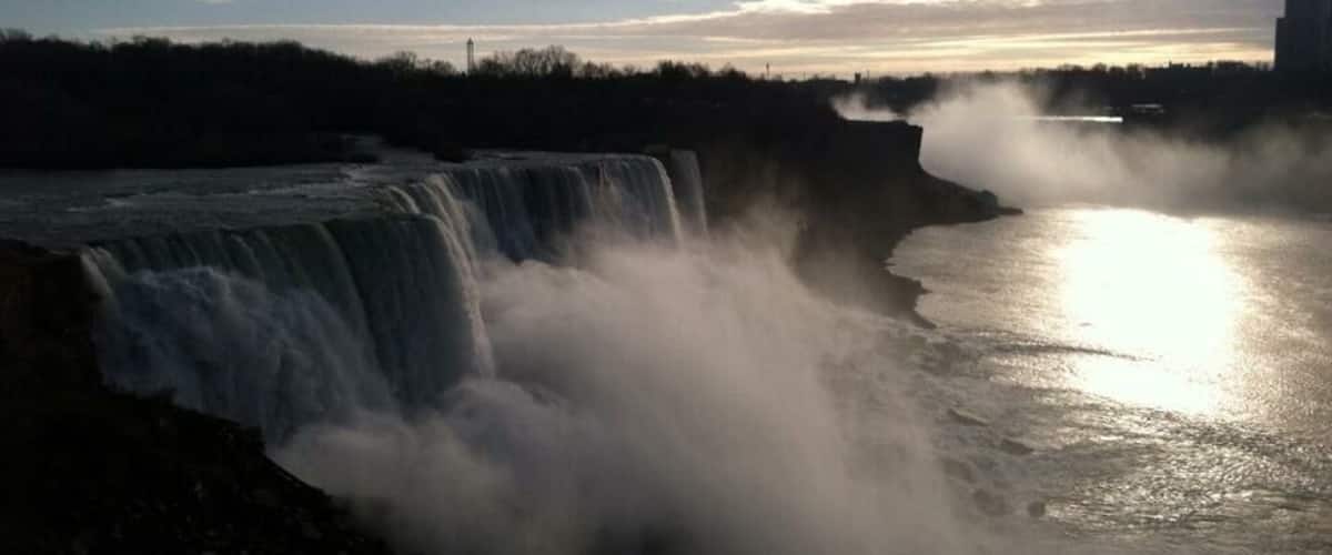 A waterfall with mist rising from the water, a clear sky, and a distant city skyline.