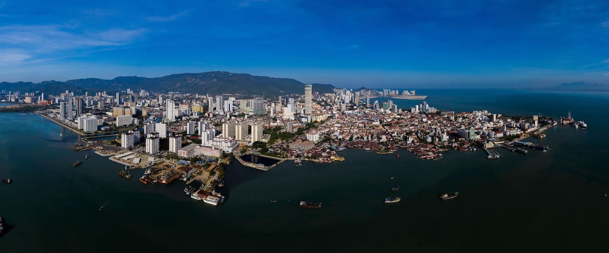 An aerial view of a coastal city with dense urban development, a prominent central island, and a backdrop of mountains.