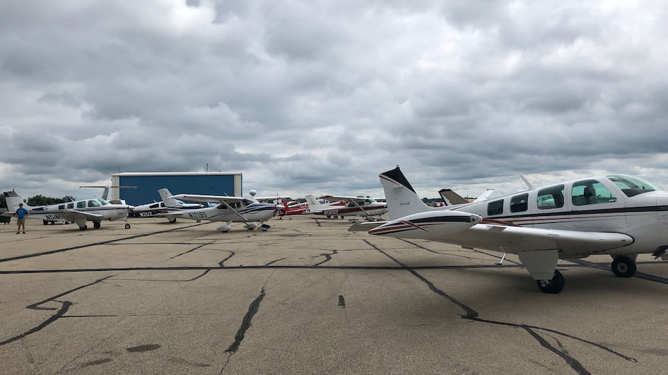 A tarmac with several small aircraft parked, a hangar in the background, and overcast skies.