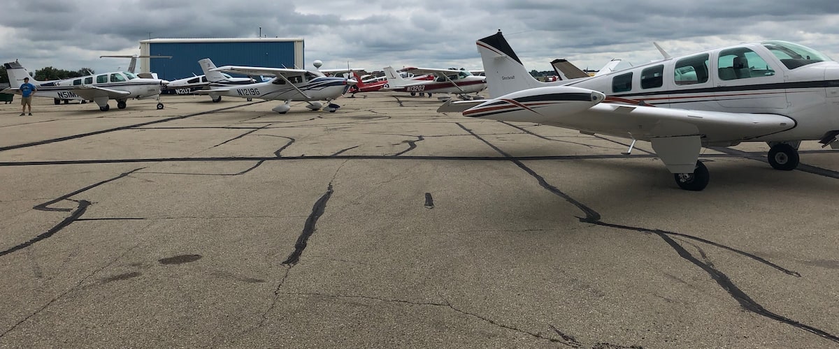 A tarmac with several small aircraft parked, a hangar in the background, and overcast skies.