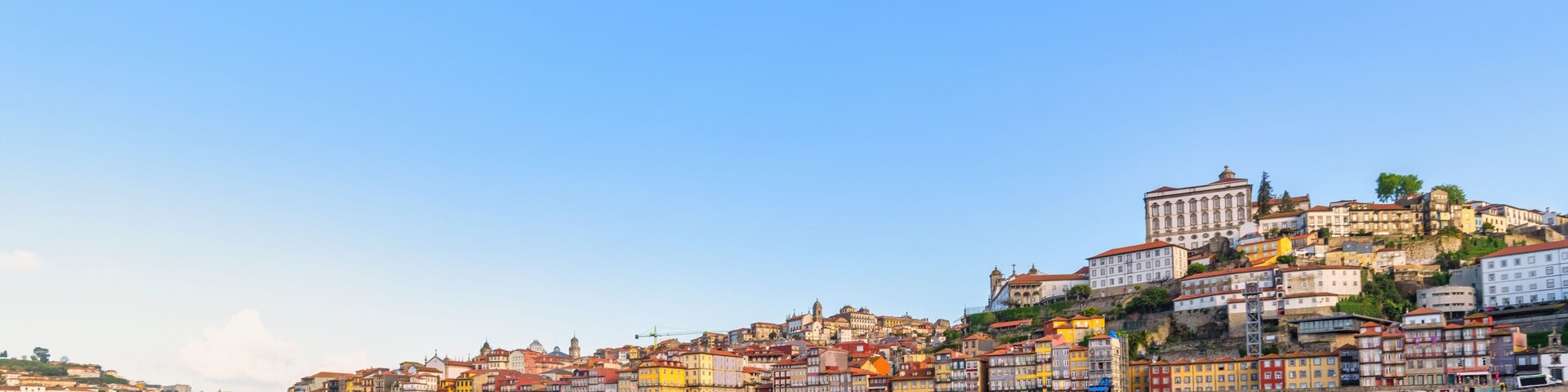 Un río que refleja edificios coloridos, una estructura destacada en una colina y un cielo azul despejado.