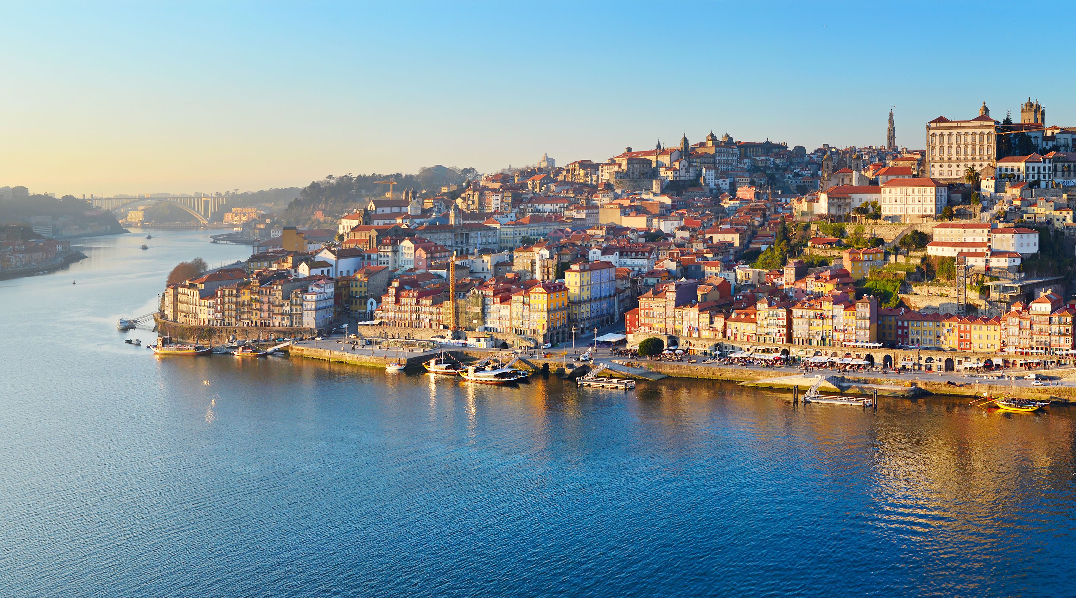 Paisagem costeira com edifícios históricos à beira-mar, uma igreja imponente e uma ponte ao longe.