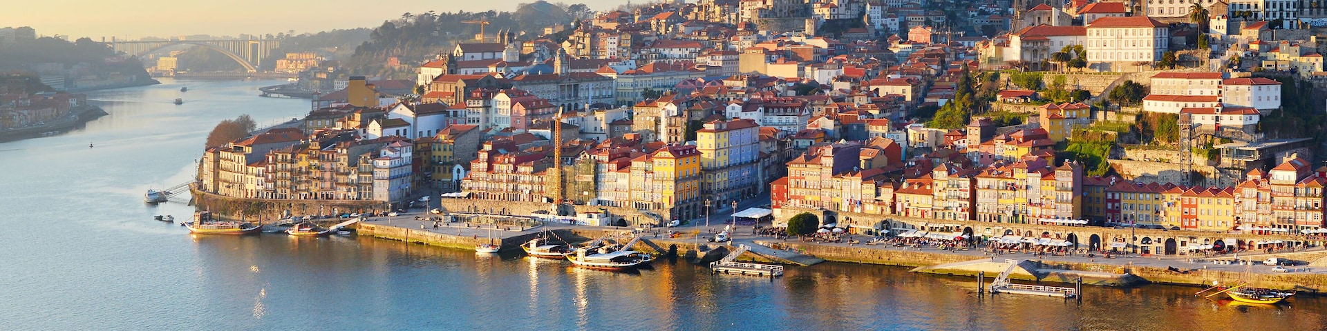 Paisagem costeira com edifícios históricos à beira-mar, uma igreja imponente e uma ponte ao longe.