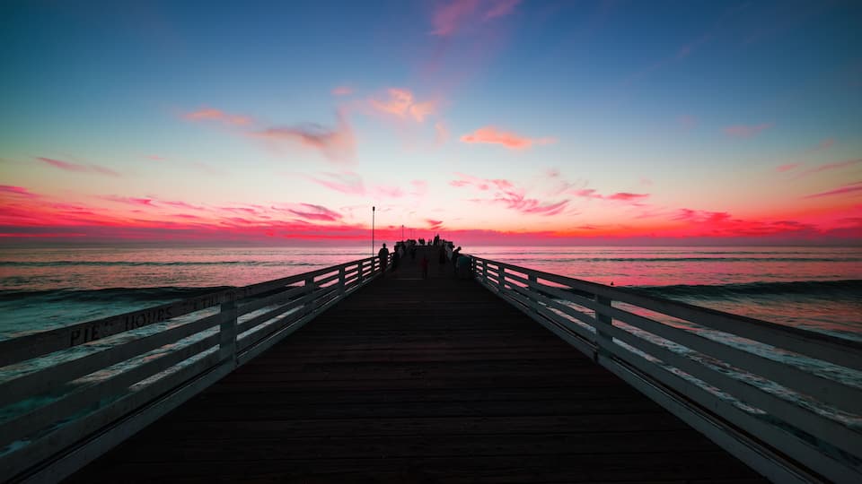 A wooden pier extending into the ocean at sunset with a pink and blue sky.
