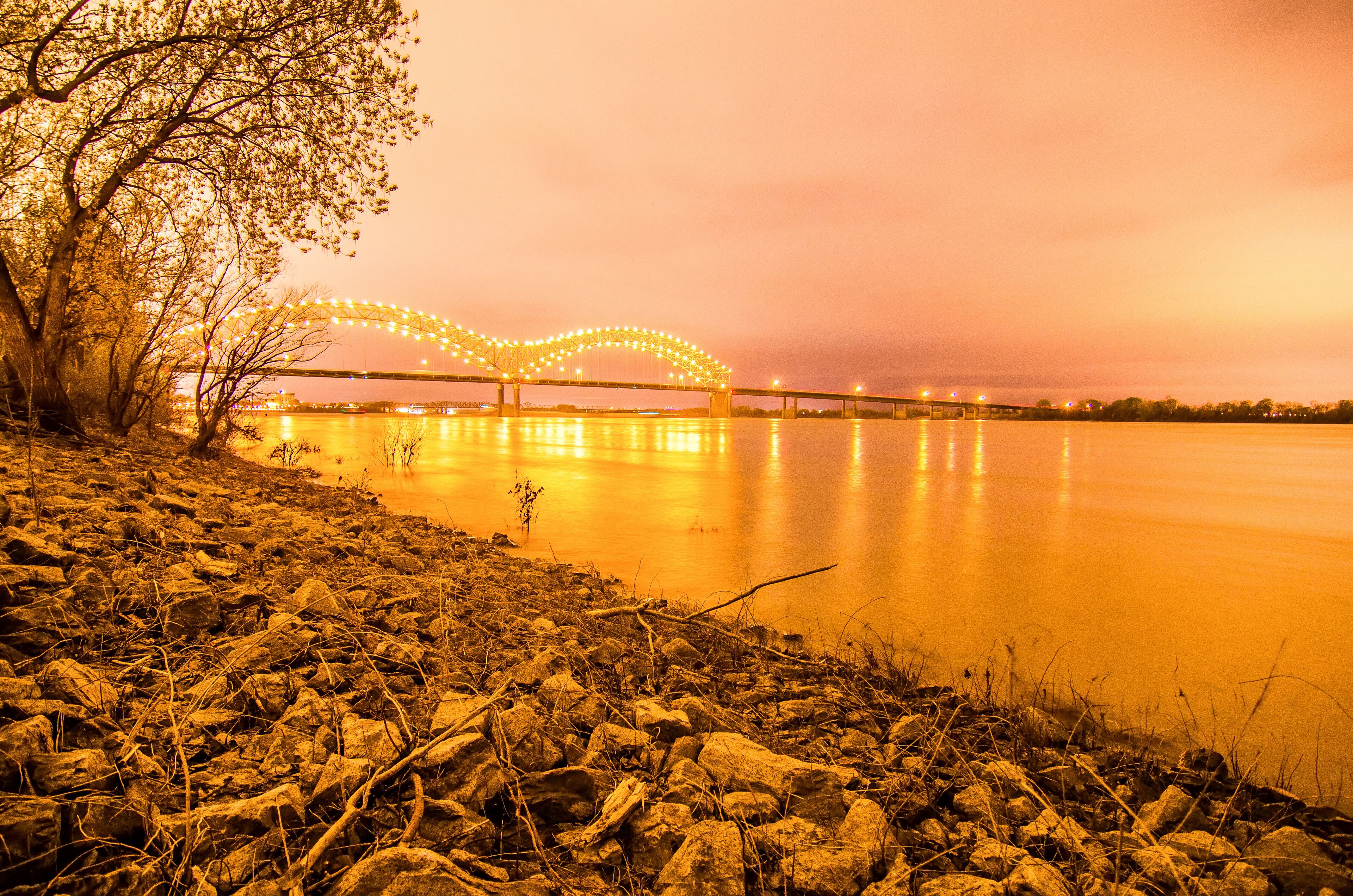 A riverbank with rocks and a tree, a bridge with lights, and a sunset sky.
