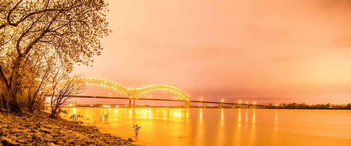 A riverbank with rocks and a tree, a bridge with lights, and a sunset sky.