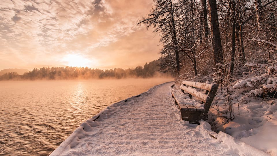 A snow-covered bench by a lake at sunset.