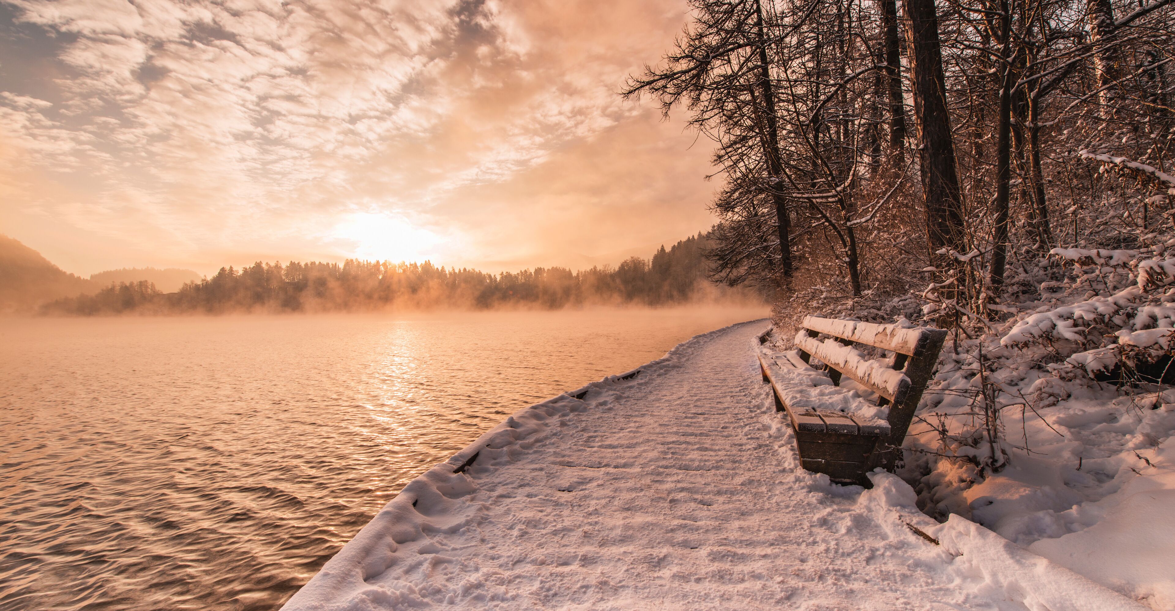 A snow-covered bench by a lake at sunset.